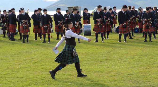 A wee lassie marches in front of a Pipe Band carrying an envelope